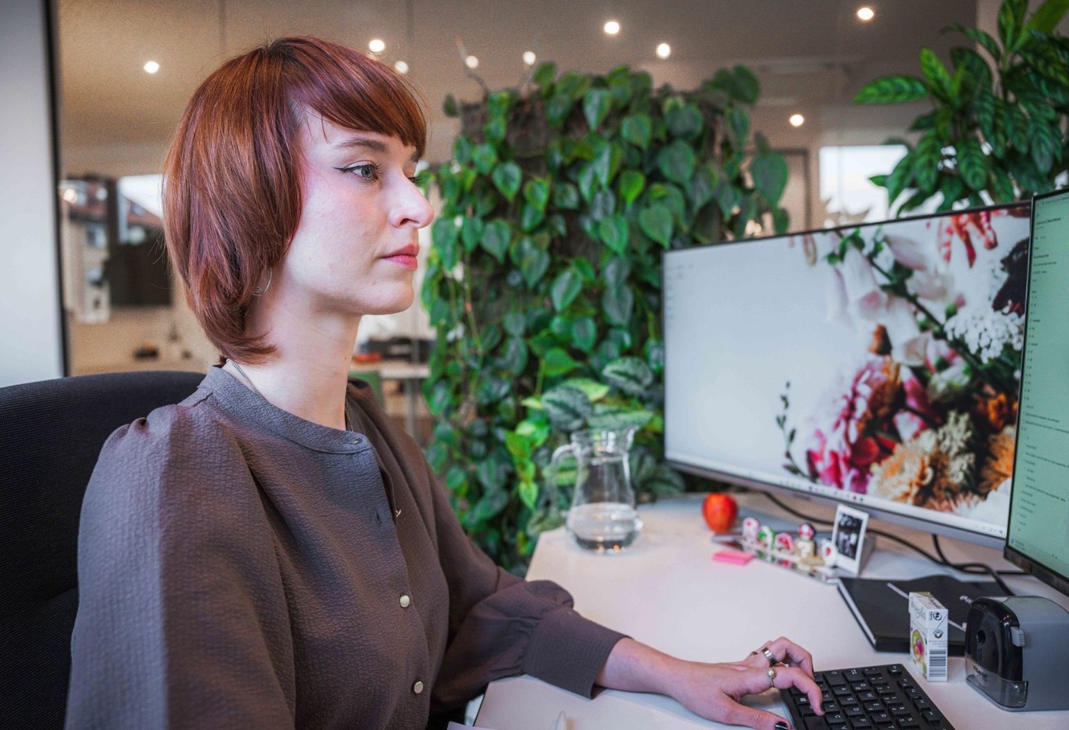 Eine Frau sitzt an einem Schreibtisch mit einem Computer und einer Pflanze im Hintergrund.
